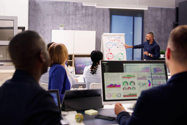 Students in a classroom with computers facing an instructor pointing to charts on a whiteboard